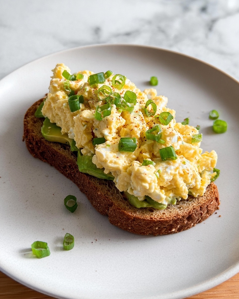 A slice of toasted brown bread sits on a plain white plate with a few green onion pieces scattered around it. On the bread is a layer of green avocado slices at the bottom, topped with a thick, chunky layer of pale yellow egg salad mixed with small white bits of egg whites. The egg salad is garnished with chopped green onions for a touch of color. The plate is on a white marbled surface, and the photo has natural lighting highlighting the textures and colors of the dish. photo taken with an iphone --ar 4:5 --v 7