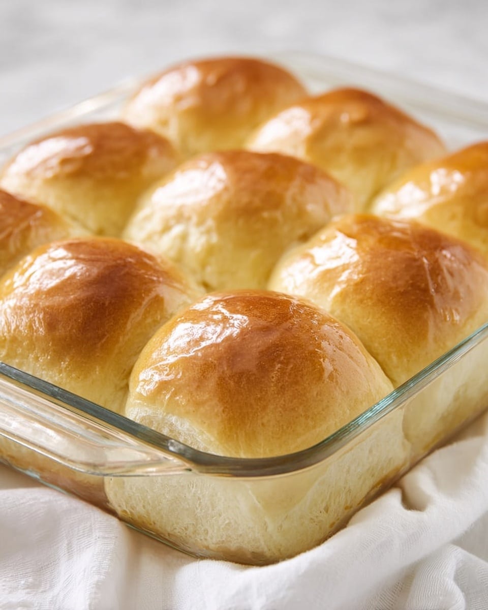 A glass baking dish filled closely with eight golden brown dinner rolls, each roll smooth on top with a shiny, slightly glazed texture, soft and fluffy in appearance. The rolls are arranged in two rows of four, all touching each other, resting on a white marbled surface with a white cloth partially visible underneath the dish. The edges of the clear dish show slight moisture, highlighting the fresh, warm condition of the rolls. Photo taken with an iphone --ar 4:5 --v 7