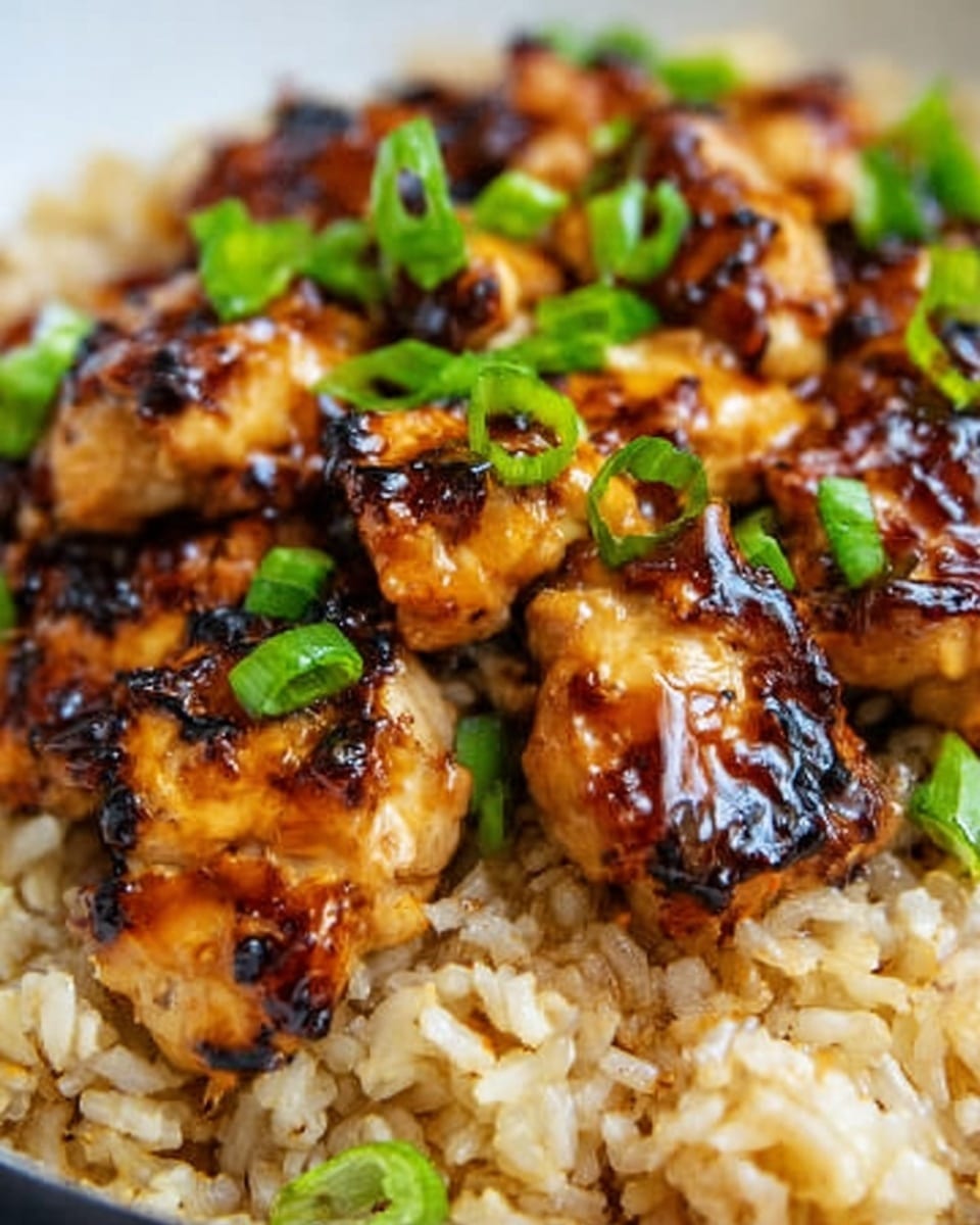 The image shows a close-up of a dish with two main layers: the bottom layer is light brown cooked rice with a soft, slightly sticky texture, and on top are pieces of grilled chicken glazed with a shiny, dark brown sauce, giving them a slightly charred look. Small bright green chopped scallions are sprinkled on the chicken, adding a fresh color contrast. The food is presented on a white plate with a white marbled surface background. photo taken with an iphone --ar 4:5 --v 7