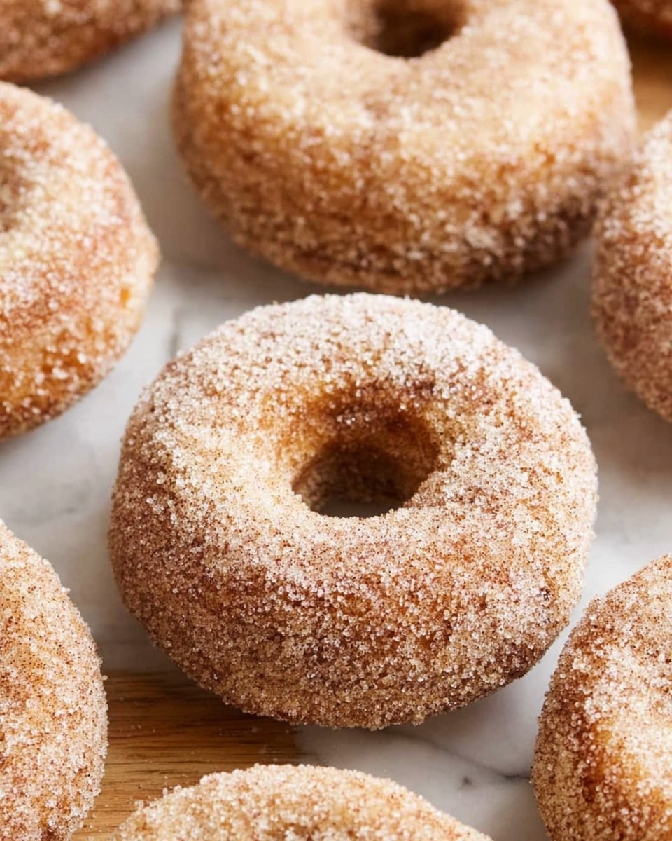 The image shows several round doughnuts with a well-defined hole in the center, each covered evenly in a layer of coarse sugar crystals mixed with cinnamon, giving a rough, textured look in light brown and white tones; the doughnuts have a golden-brown color showing through the sugar-coating, and they are arranged closely on a white marbled surface, creating a warm and inviting atmosphere. photo taken with an iphone --ar 4:5 --v 7