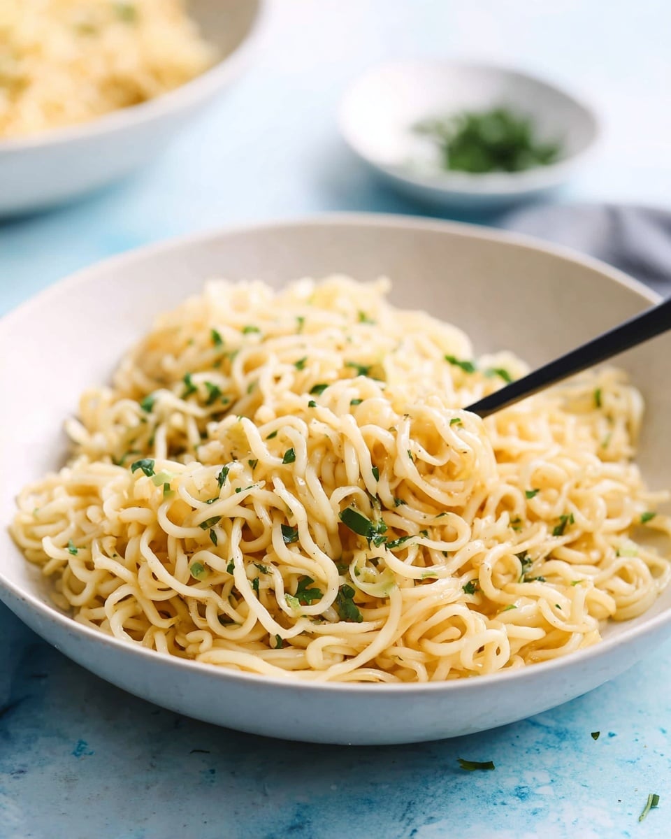 A close-up view of a shallow white bowl filled with curly cooked noodles that have a light yellow color, sprinkled lightly with green chopped herbs. The noodles form a single big mound with soft, slightly shiny texture, and some herbs are scattered throughout the top layer, adding green contrast. A black fork is partially stuck into the noodles, positioned on the right side. The bowl is placed on a white marbled surface with a soft blue hue in the background, showing slight blur and a hint of another white bowl and a small dish with more herbs in the background. photo taken with an iphone --ar 4:5 --v 7