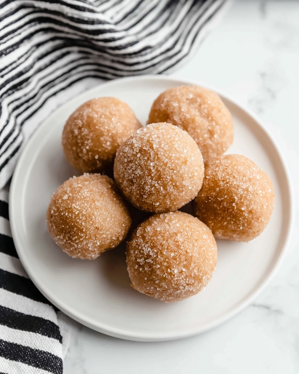 Seven round, light brown dough balls are placed closely together on a white plate. Each ball has a rough texture with coarse sugar crystals sprinkled evenly over the surface, giving them a sparkly, crunchy look. The plate is set on a white marbled surface with a black and white striped cloth visible in the background. The dough balls appear soft but firm, arranged in a simple cluster. photo taken with an iphone --ar 4:5 --v 7