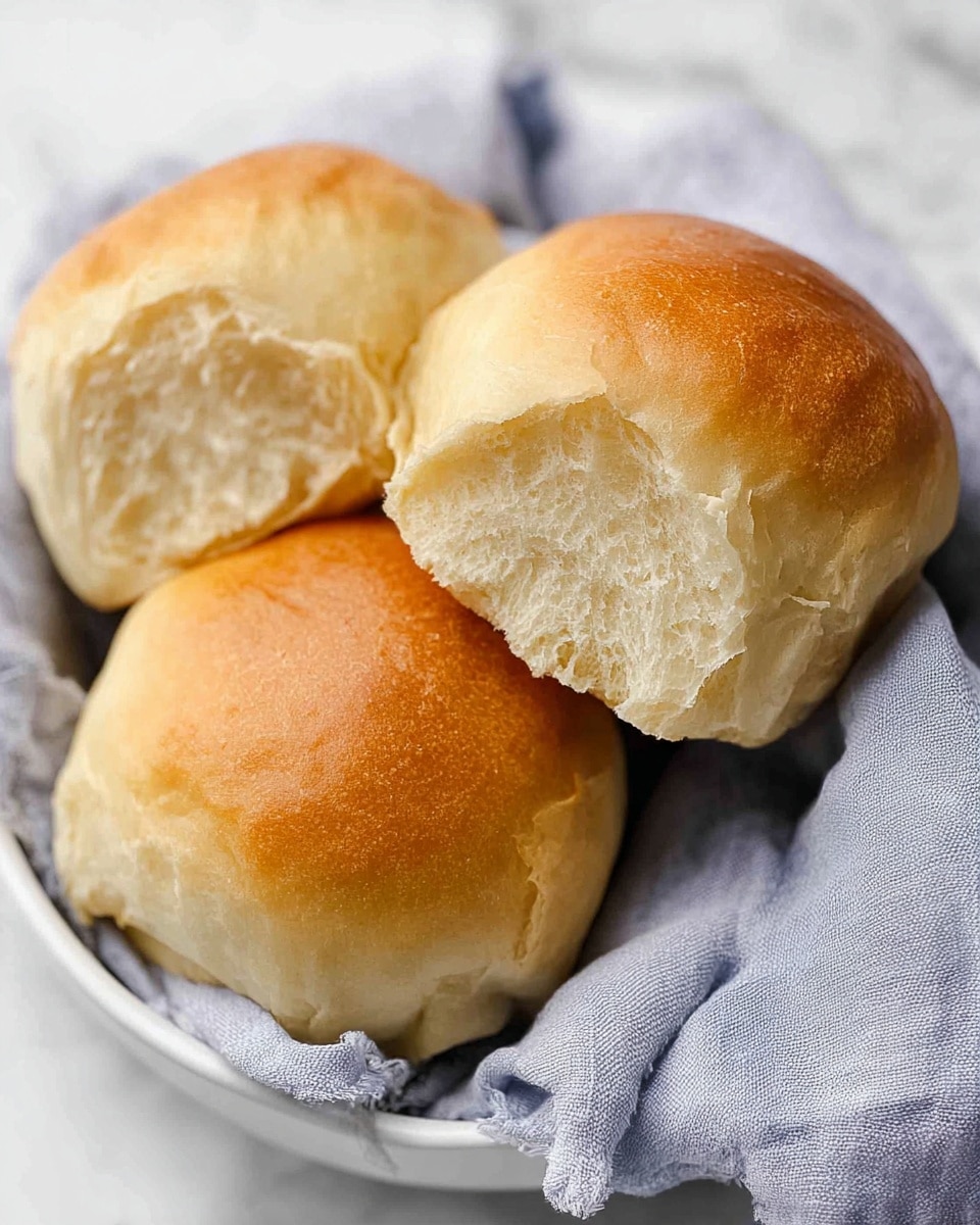 The image shows three soft, golden-brown dinner rolls nestled together in a white bowl lined with a light gray cloth. Each roll has a smooth, slightly shiny top with a fluffy, light interior visible in the torn part of one roll. The rolls appear warm and fresh, with a gentle texture that looks tender and airy. The white bowl and the gray cloth create a calm and simple setting, all set on a white marbled surface. photo taken with an iphone --ar 4:5 --v 7