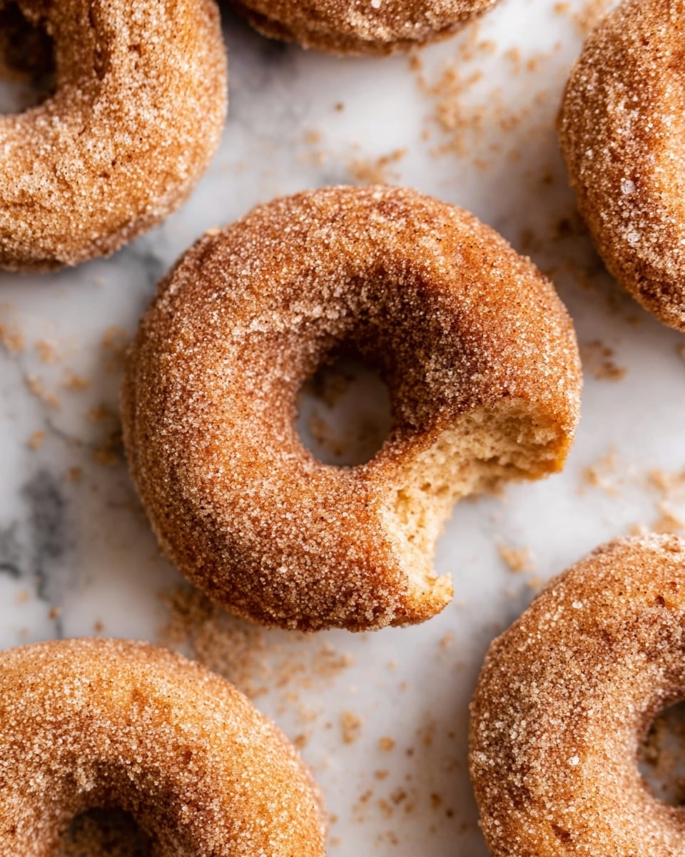 The image shows several cinnamon sugar-coated donuts with a light brown color and a rough sugary texture covering their surface. One donut is in the center with a bite taken out of it, revealing a soft, crumbly inside with a light golden hue. The donuts have a classic ring shape and are placed on a white marbled surface with some sugar crumbs scattered around. The overall look is warm and inviting, highlighting the crunchy sugar layer and the soft dough inside. photo taken with an iphone --ar 4:5 --v 7