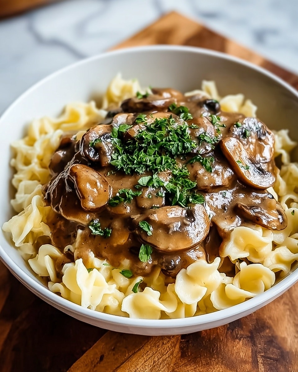 A white bowl holds a creamy mushroom pasta dish with two main layers; at the bottom is soft, pale yellow pasta with a smooth, slightly glossy texture, topped with a generous serving of rich brown mushroom sauce that includes sliced mushrooms cooked in a thick sauce. The dish is finished with a sprinkle of bright green chopped herbs on top, adding a fresh contrast. The bowl is placed on a wooden surface with a white marbled texture underneath. Photo taken with an iphone --ar 4:5 --v 7