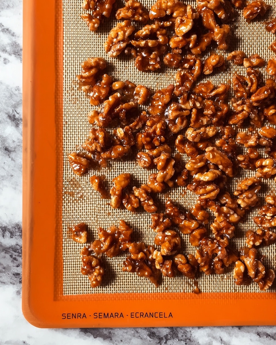 The image shows a single flat layer of shiny, candied walnut pieces spread evenly on a beige baking mat with an orange border placed on a white marbled texture. The walnuts have a rich, glossy, dark brown coating that looks sticky and caramelized, giving them a textured and slightly irregular shape. The pieces vary in size, some larger clusters mixed with smaller bits, all glistening under soft light. photo taken with an iphone --ar 4:5 --v 7