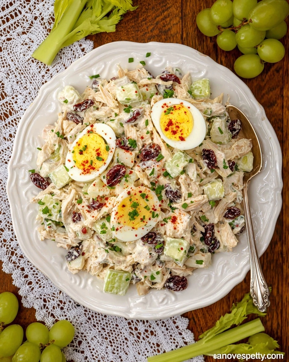 A white decorative plate holds a creamy chicken salad with visible shredded white chicken, light green grape halves, celery pieces, and bits of dark dried cranberries mixed throughout. The salad is topped with two halved hard-boiled eggs, each showing a bright yellow yolk sprinkled with red paprika and finely chopped green herbs. The plate sits on a wooden surface with a white lace cloth partially visible, along with loose green grapes and celery sticks around it. A large silver spoon is placed next to the plate. photo taken with an iphone --ar 4:5 --v 7