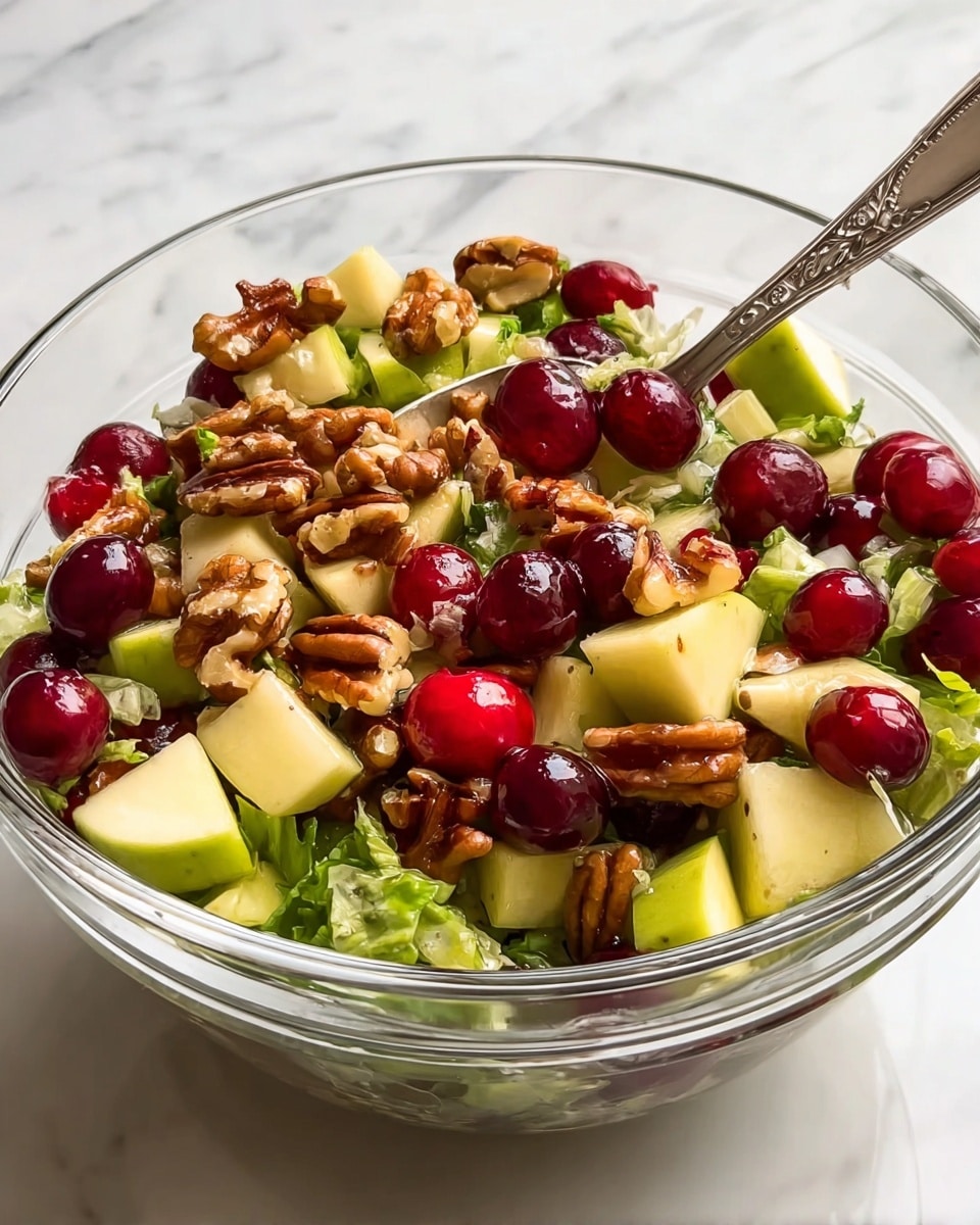 A clear glass bowl filled with a colorful salad showing multiple layers: the bottom layer consists of chopped green lettuce pieces, above it are light yellow chunks of apple mixed with fresh green apple slices, then bright red cranberries and dark red grapes are scattered throughout, and the top layer is covered with large pieces of shelled walnuts. A silver spoon rests inside the bowl, partially submerged in the salad and shiny, with a few cranberries sitting on it. The bowl is set on a white marbled surface. photo taken with an iphone --ar 4:5 --v 7