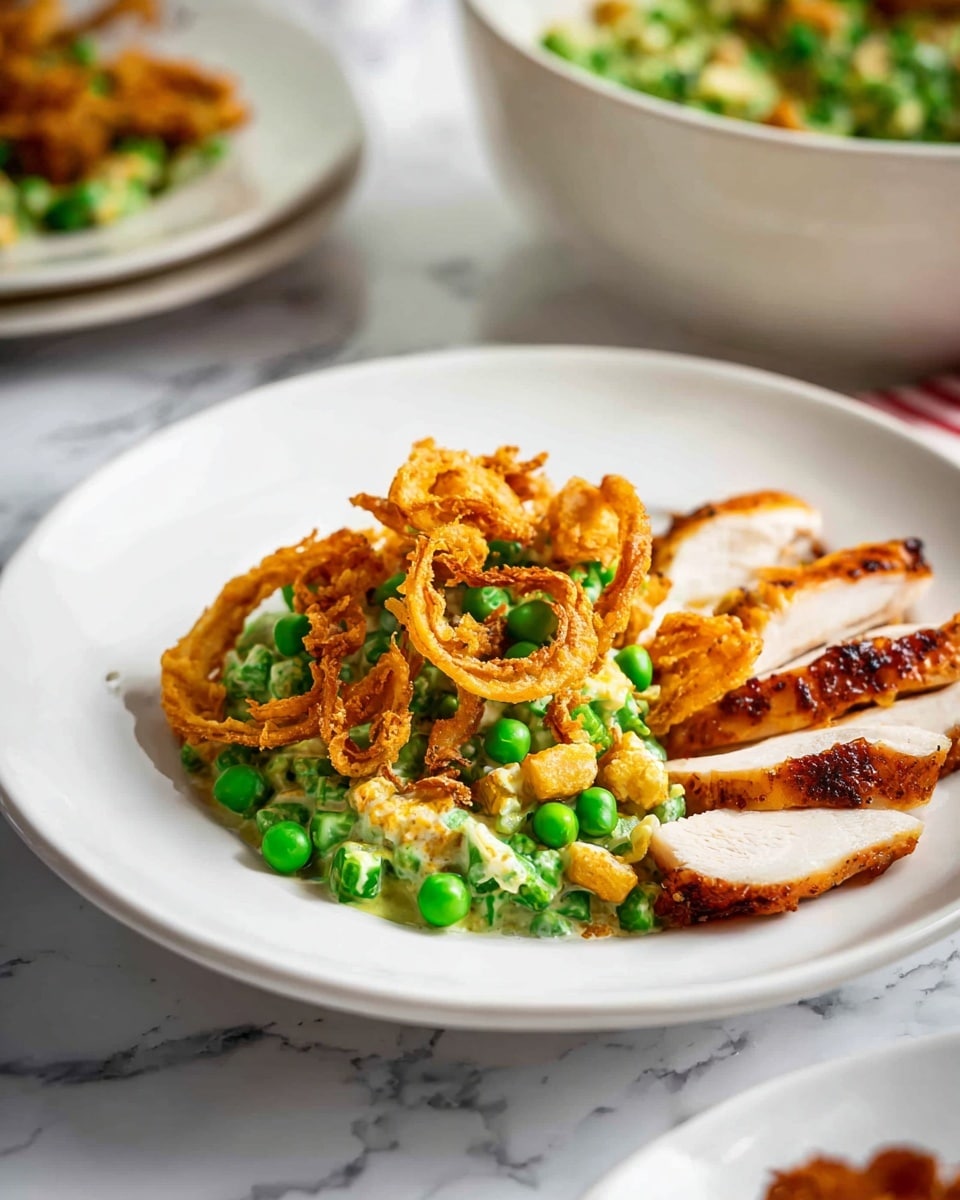 The dish is served on a white plate placed on a white marbled surface. The main section of the plate holds a creamy green pea salad mixed with crispy fried onion rings on top, creating a mix of vibrant green and golden-brown colors with a creamy texture underneath. To the side, there are three thin slices of roasted chicken breast with a golden-brown, slightly charred crust, showing juicy white meat inside. In the background, a similar white plate and a white bowl with more of the pea salad are partially visible, adding depth to the scene. photo taken with an iphone --ar 4:5 --v 7