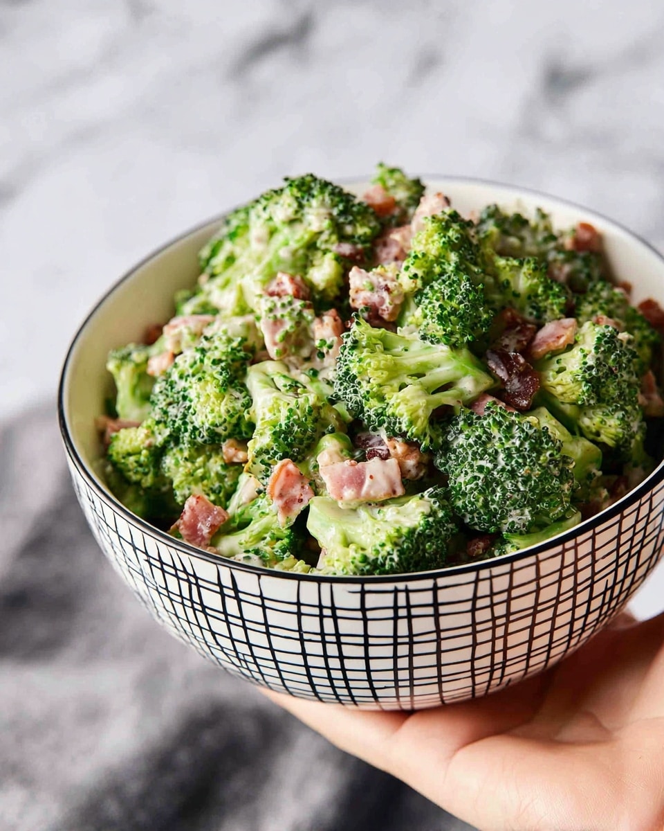 A close-up of a white bowl with black square patterns filled with a broccoli salad. The salad has three main layers: bright green broccoli florets with a bumpy texture, small pieces of pinkish bacon scattered throughout, and creamy white dressing coating the ingredients lightly. The bowl is held by a woman's hand at the bottom right corner, all placed against a white marbled background. photo taken with an iphone --ar 4:5 --v 7