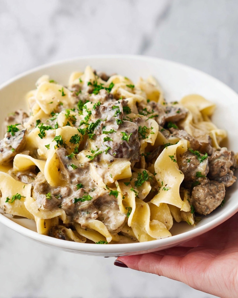 A white bowl filled with a creamy beef stroganoff dish is shown, held by a woman's hand at the bowl's edge. Inside, there are wide, flat egg noodles with a pale yellow color, mixed with pieces of ground beef and sliced mushrooms coated in a thick, light brown sauce. Small bits of bright green parsley are sprinkled on top, adding color contrast. The background is a white marbled texture. photo taken with an iphone --ar 4:5 --v 7