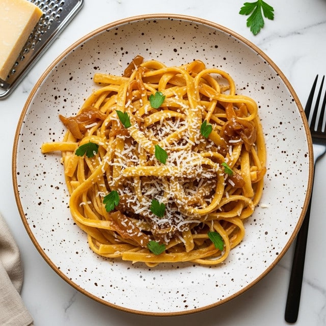 A white speckled plate contains a serving of linguine pasta coated in a creamy light brown sauce with visible soft caramelized onion slices mixed throughout. The pasta strands, pale yellow in color, are loosely twirled in the center of the plate, sprinkled with finely chopped green parsley and tiny shreds of white grated cheese. Small black pepper flakes are scattered on top, adding contrast to the dish. Next to the plate, on a white marbled surface, there is a block of hard cheese with some grating near a metal grater, and a black fork is placed on the right side. photo taken with an iphone --ar 4:5 --v 7