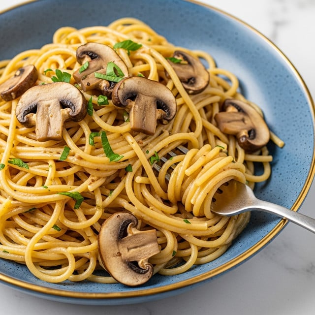 A close-up of a serving of spaghetti pasta mixed with light brown creamy sauce, scattered with sliced brown mushrooms and small pieces of fresh green parsley on top. The pasta strands are smooth and glossy, some twirled around a silver fork resting in the blue bowl. The bowl has a speckled texture and a golden rim, placed on a white marbled surface. photo taken with an iphone --ar 4:5 --v 7