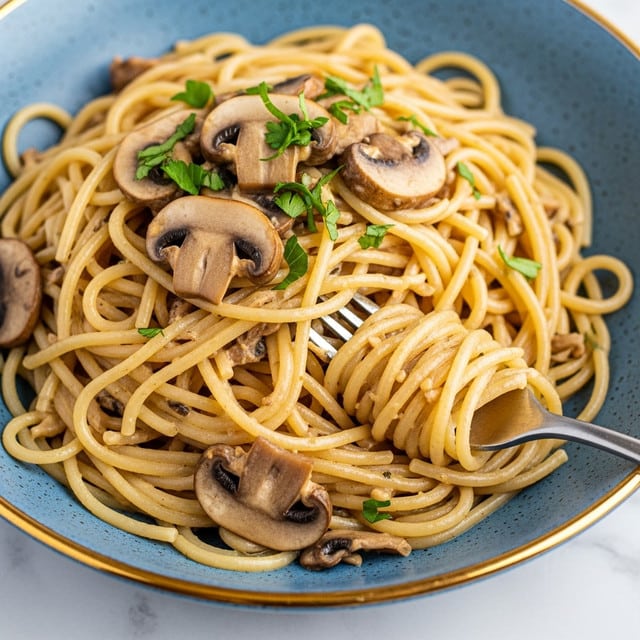 A close-up view of a pasta dish showing long, smooth, light golden spaghetti noodles mixed with sliced brown mushrooms scattered throughout. Bright green sprinkled parsley pieces add color contrast on top. The pasta is glossy, indicating it is coated in a creamy sauce. A silver fork is twirling some noodles with a couple of mushroom slices held above the plate. The white plate has a slightly raised edge and the background is a white marbled texture. photo taken with an iphone --ar 4:5 --v 7