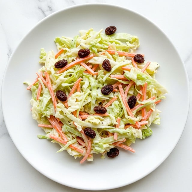 A white plate holds a single layer of creamy coleslaw, made of shredded pale green cabbage mixed with thin orange carrot strips and scattered dark raisins. The textures of the cabbage and carrots contrast with the smooth, creamy dressing coating the vegetables. The coleslaw is piled unevenly, giving a fresh and slightly messy look. The plate sits on a white marbled surface. photo taken with an iphone --ar 4:5 --v 7