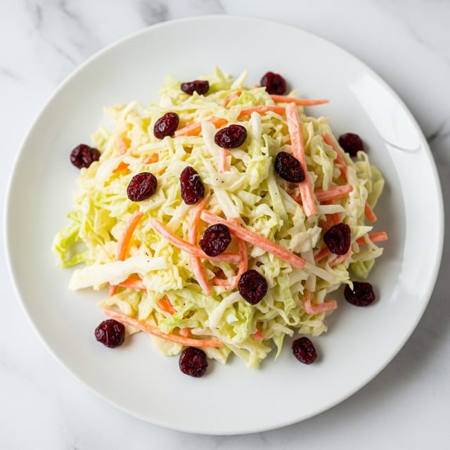 A white plate holds a mound of coleslaw made of three visible layers: shredded pale green cabbage forming the base with a rough texture, mixed with thin orange carrot strips evenly spread throughout, topped with a creamy white dressing that coats all ingredients giving a smooth and moist look, scattered with small dark red dried cranberries adding contrast and color spots all around. The plate sits on a white marbled texture. Photo taken with an iphone --ar 4:5 --v 7