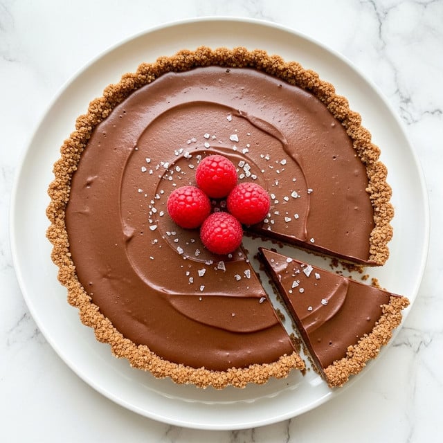 A tart with two main layers is shown on a white plate against a white marbled textured surface. The bottom crust layer is light brown with a crumbly texture, and the top layer is a smooth, glossy dark chocolate filling. The tart is sliced, with two slices slightly separated from the whole. Three bright red raspberries are placed in the center on top, with small white salt flakes sprinkled around the surface. Photo taken with an iphone --ar 4:5 --v 7