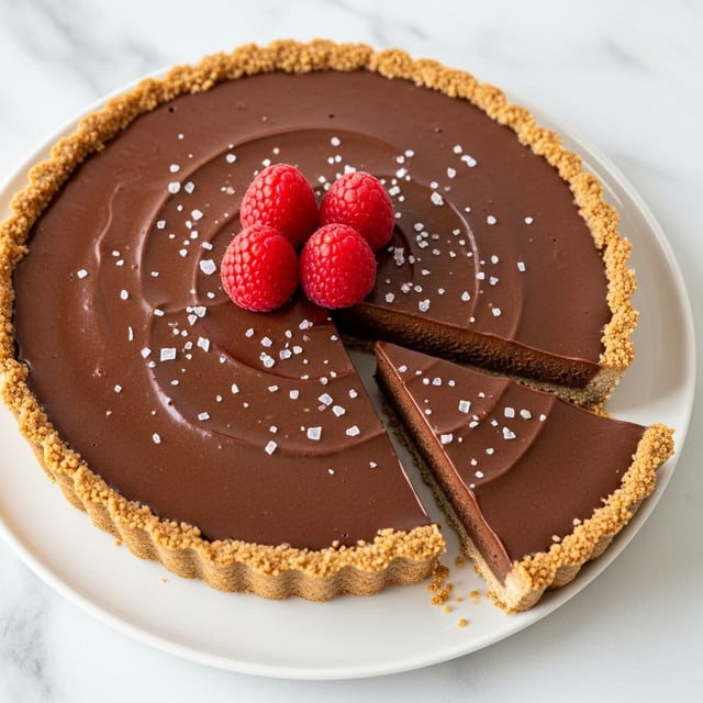 A chocolate tart with a smooth, shiny dark brown chocolate layer on top is cut into slices, showing a thick, slightly textured chocolate filling above a golden-brown, crumbly crust at the bottom. Three bright red raspberries sit in the center of the tart, and small white flakes, probably sea salt, are scattered on the chocolate surface. The tart rests on a white plate placed on a white marbled surface. Photo taken with an iphone --ar 4:5 --v 7
