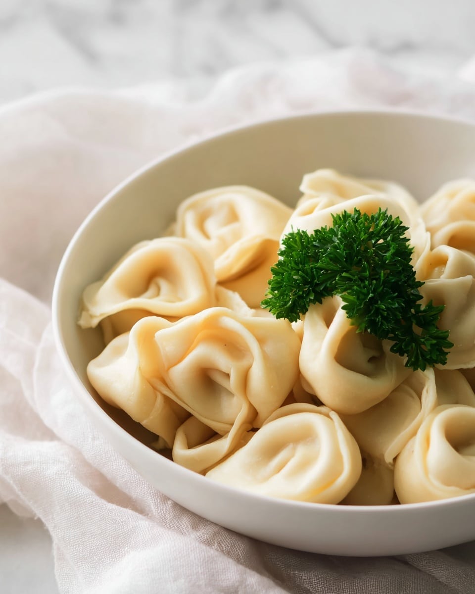 The image shows a bowl filled with many pieces of tortellini pasta. The pasta is pale yellow with a soft, cooked texture, each piece folded neatly into the classic ring shape. On top of the pasta is one small green sprig of parsley for decoration. The bowl is white and round, placed on a white marbled surface with some blurred green plants in the background. Photo taken with an iphone --ar 4:5 --v 7