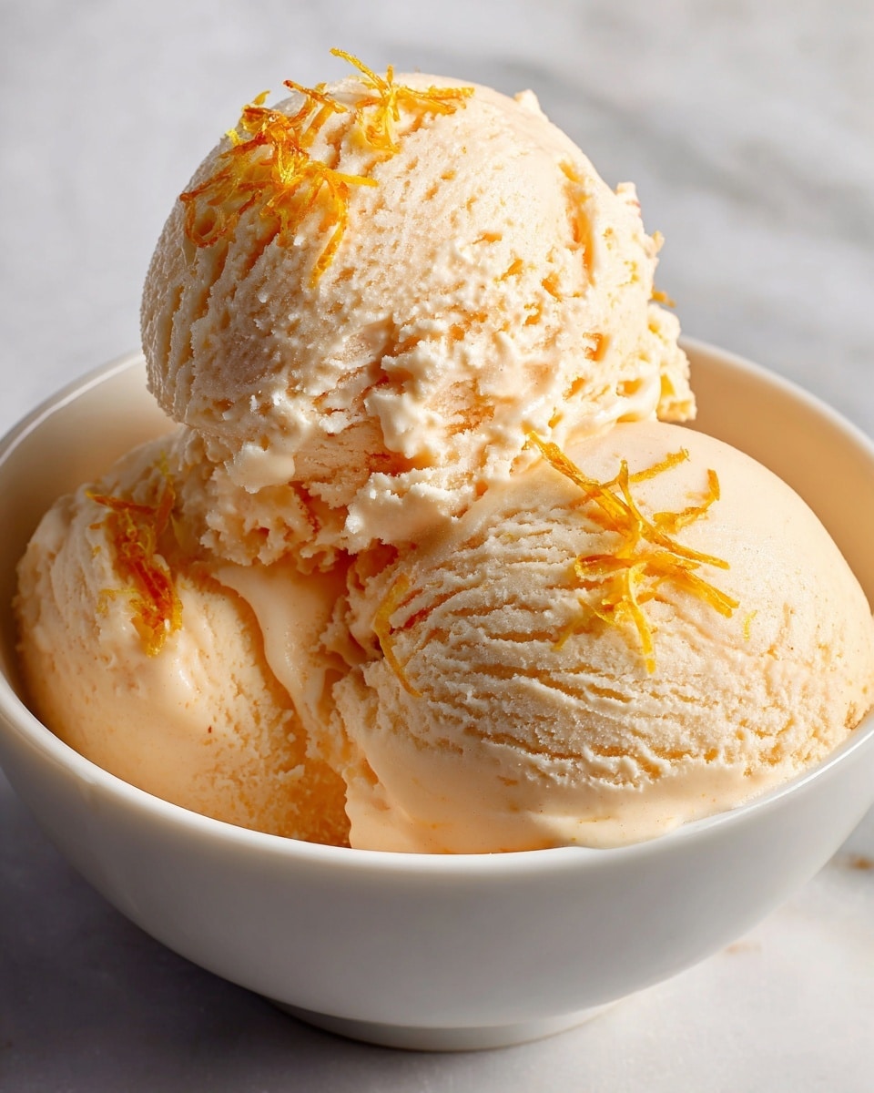 A close-up image of three ice cream scoops in a white bowl set on a white marbled surface. The ice cream is a creamy orange color with a smooth but slightly textured surface showing soft ridges and small air pockets. The top scoop is sprinkled with small bright orange zest shreds that add texture and visual contrast to the smooth ice cream beneath. Soft shadows and warm lighting highlight the shiny, slightly wet look on parts of the ice cream, giving it a fresh and appetizing appearance. photo taken with an iphone --ar 4:5 --v 7