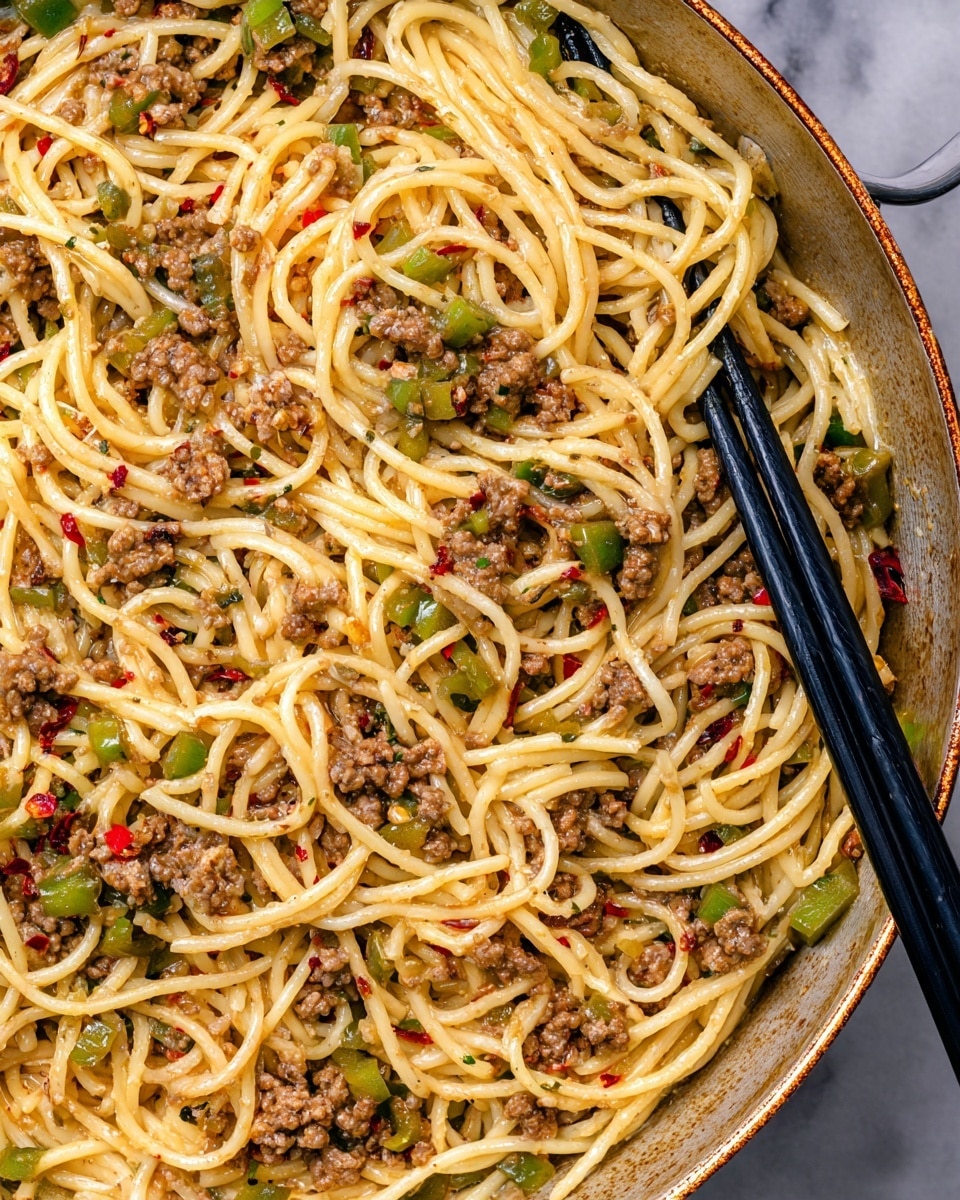 The image shows a close-up of a large white pan filled with spaghetti mixed with small pieces of cooked ground meat and chopped green bell peppers. The spaghetti is yellow and shiny, coated in a light sauce with hints of red chili flakes scattered throughout, adding specks of red color. The ground meat is browned and crumbly, evenly spread among the noodles. Two black chopsticks rest inside the pan, positioned on the right side, slightly mixing the pasta. The background surface is a white marbled texture. photo taken with an iphone --ar 4:5 --v 7