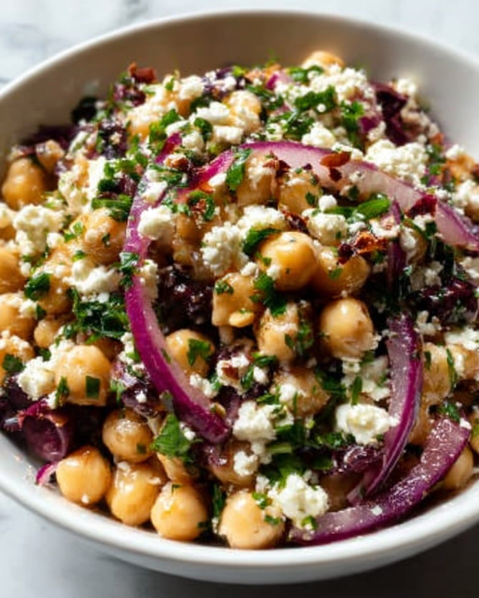A close-up view of a white bowl filled with a layered salad composed of round beige chickpeas, soft crumbled white cheese, bright green chopped herbs, and slices of purple-red onions. The chickpeas create the base layer mixed with the thinly sliced onions, while the white cheese pieces and green herbs are sprinkled on top. There are also small bits of a dark ingredient, possibly olives or dried fruit, scattered throughout the bowl. The bowl is placed on a white marbled surface, and the textures show a mix of smooth chickpeas, crumbly cheese, and slightly shiny onion slices. The lighting gives a fresh, natural look. Photo taken with an iphone --ar 4:5 --v 7
