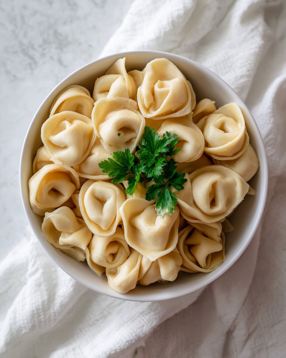A close-up view of a round white bowl filled with many light beige tortellini pasta pieces, each folded into a ring shape with smooth texture and slightly glossy surface. The pasta is stacked naturally with some pieces leaning on others, creating depth. In the center, there is a small bunch of fresh dark green parsley leaves on top, adding a touch of color contrast. The bowl sits on a soft white cloth with subtle creases, set against a background of white marbled texture. Photo taken with an iphone --ar 4:5 --v 7