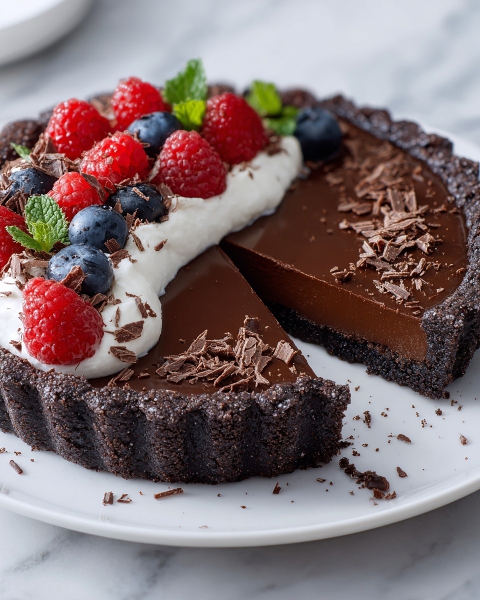 A dark chocolate tart on a white plate is shown, with one slice removed. The tart has three visible layers: a crumbly black crust on the bottom and sides, a smooth and shiny dark chocolate filling in the middle, and a layer of white whipped cream topped with fresh red raspberries and blue blueberries on one side. Small dark chocolate shavings add texture, scattered on the whipped cream and around the edge of the tart. The plate is set on a white marbled surface. In the background, a white bowl filled with more chocolate shavings is slightly out of focus. Photo taken with an iphone --ar 4:5 --v 7
