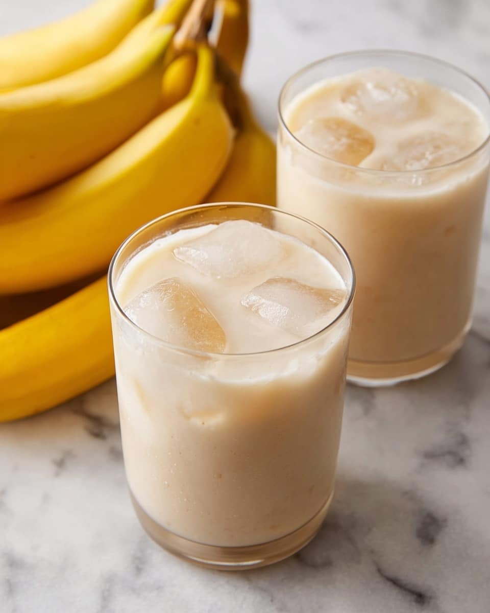 The image shows two clear glasses filled with a creamy light beige banana smoothie. Each glass contains some ice cubes visible through the liquid. In the background, there is a bunch of ripe yellow bananas resting on a white marbled surface. The smoothie has a smooth, frothy texture on top and the glasses are simple and round in shape. photo taken with an iphone --ar 4:5 --v 7
