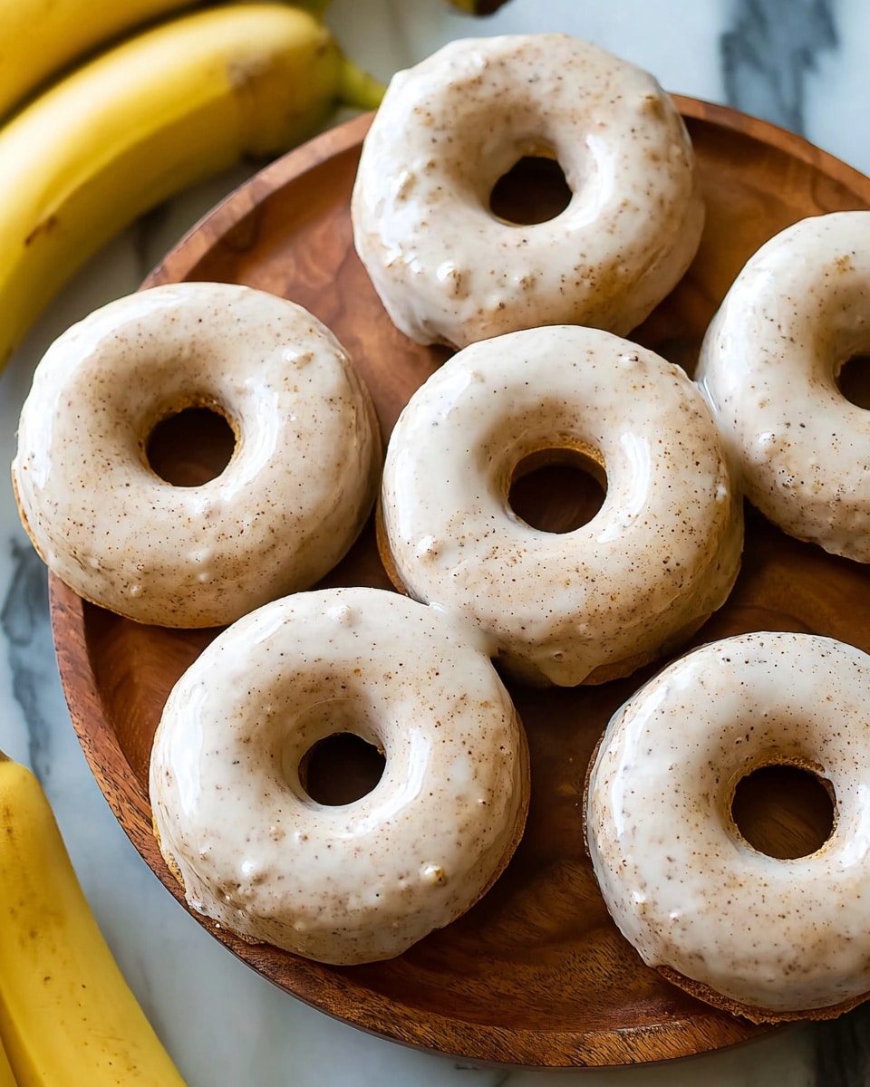 Five light brown donuts with a smooth, shiny glaze covering them evenly are arranged on a round white wooden board. The donuts have a soft, spongy texture with small specks visible under the glaze. In the background, a few ripe yellow bananas rest slightly out of focus. The whole scene is set on a white marbled surface. Photo taken with an iphone --ar 4:5 --v 7