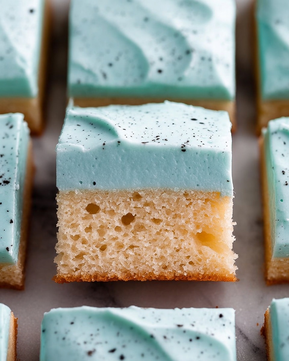 The image shows a close-up of a slice of cake resting in a larger tray of square cake pieces. The cake has two layers: the bottom layer is a moist, light brown sponge cake with a soft texture, and the top layer is a thick, smooth pale blue-green frosting sprinkled with small dark specks. The slice appears to be freshly cut, with sharp edges and the surrounding square pieces matching the same frosting and texture. The background is a white marbled texture. Photo taken with an iphone --ar 4:5 --v 7