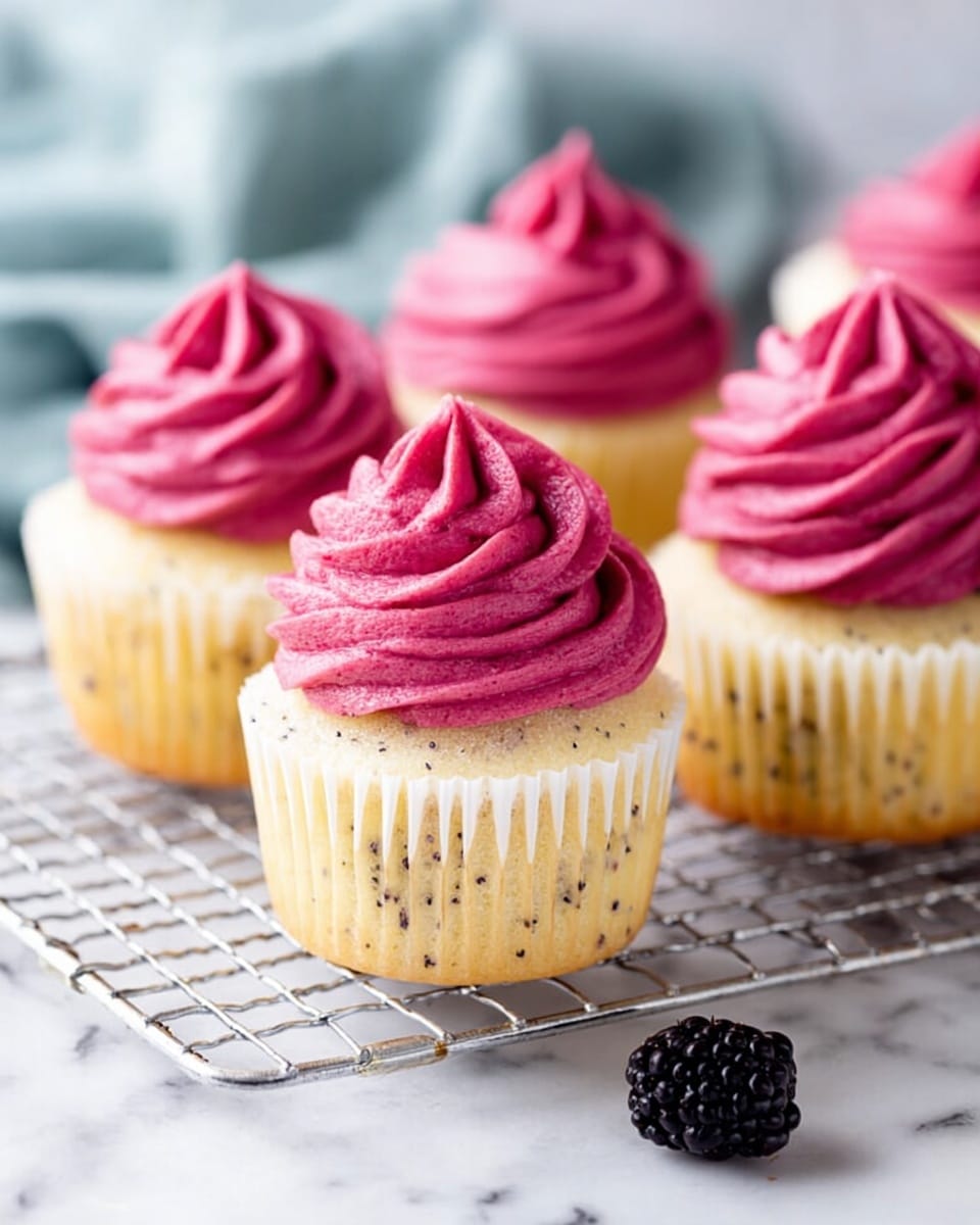 The image shows five vanilla cupcakes with poppy seeds, each topped with a thick layer of smooth, bright pink frosting swirled in a pointed, curly shape. The cupcakes sit on a silver wire cooling rack placed over white parchment paper on a white marbled surface. In the bottom right corner, a small group of shiny blackberries rests next to the rack. The background is softly blurred, emphasizing the cupcakes in the front. photo taken with an iphone --ar 4:5 --v 7