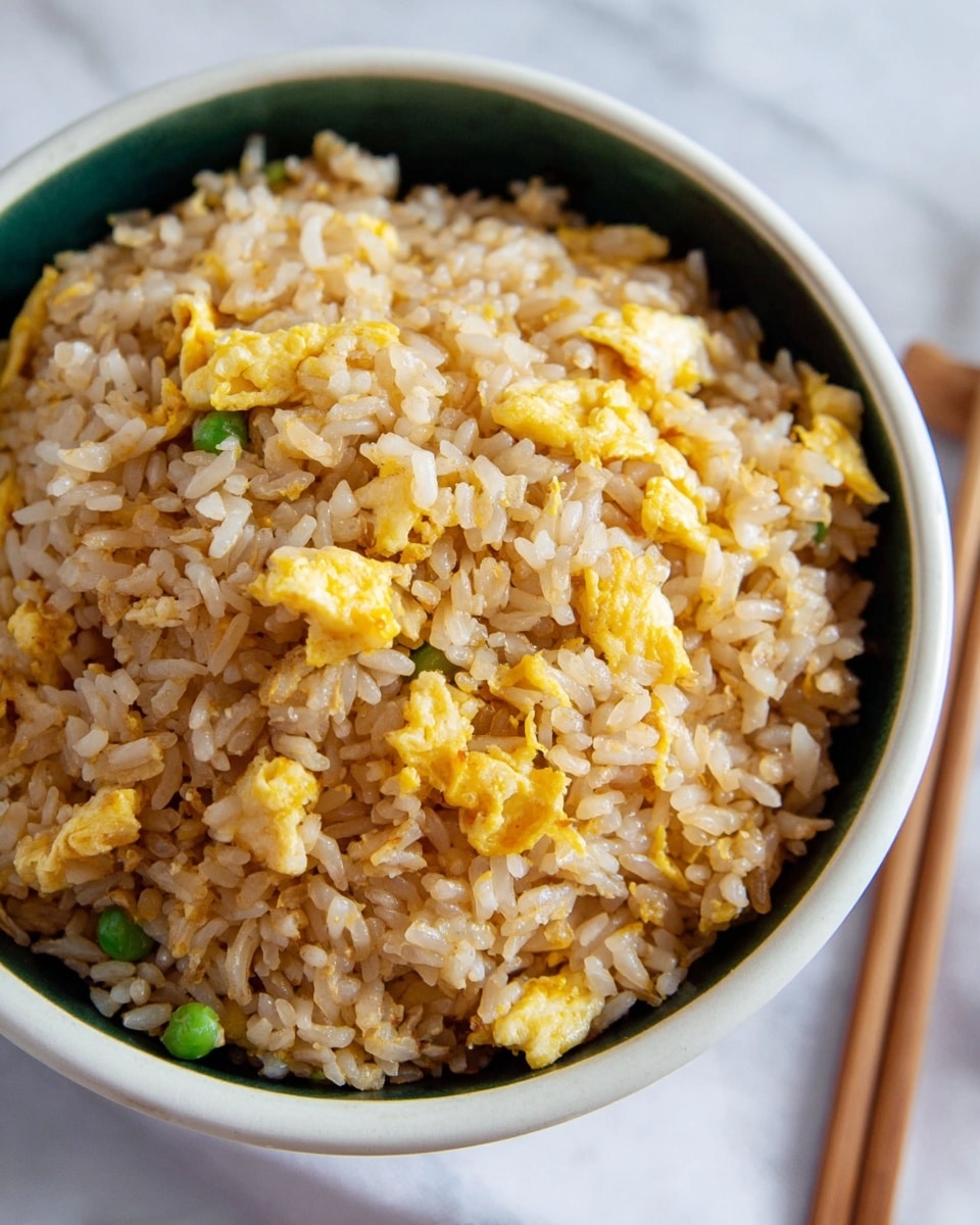 A close-up of a bowl filled with fried rice mixed with scrambled egg pieces. The dish has two main layers: the rice grains, which are light brown and slightly shiny, form the base, and scattered throughout are small, bright yellow scrambled egg chunks with a soft texture. The bowl is white on the outside and dark green on the inside, contrasting with the rice. The surface underneath is a white marbled texture with a soft natural light coming from the side. photo taken with an iphone --ar 4:5 --v 7
