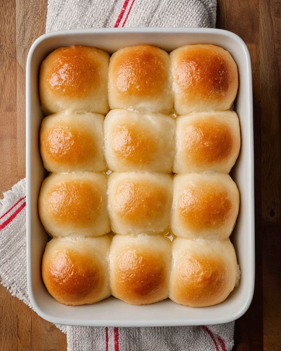 A white rectangular baking dish holds 12 soft, golden-brown dinner rolls arranged in a 3 by 4 grid. Each roll has a smooth, slightly shiny surface with a light golden top that fades to a pale beige on the sides, showing a fluffy texture. The rolls are close together, touching each other, and look freshly baked. The dish sits on a wooden table with a white cloth towel that has red and gray stripes partially visible underneath the dish. Photo taken with an iphone --ar 4:5 --v 7