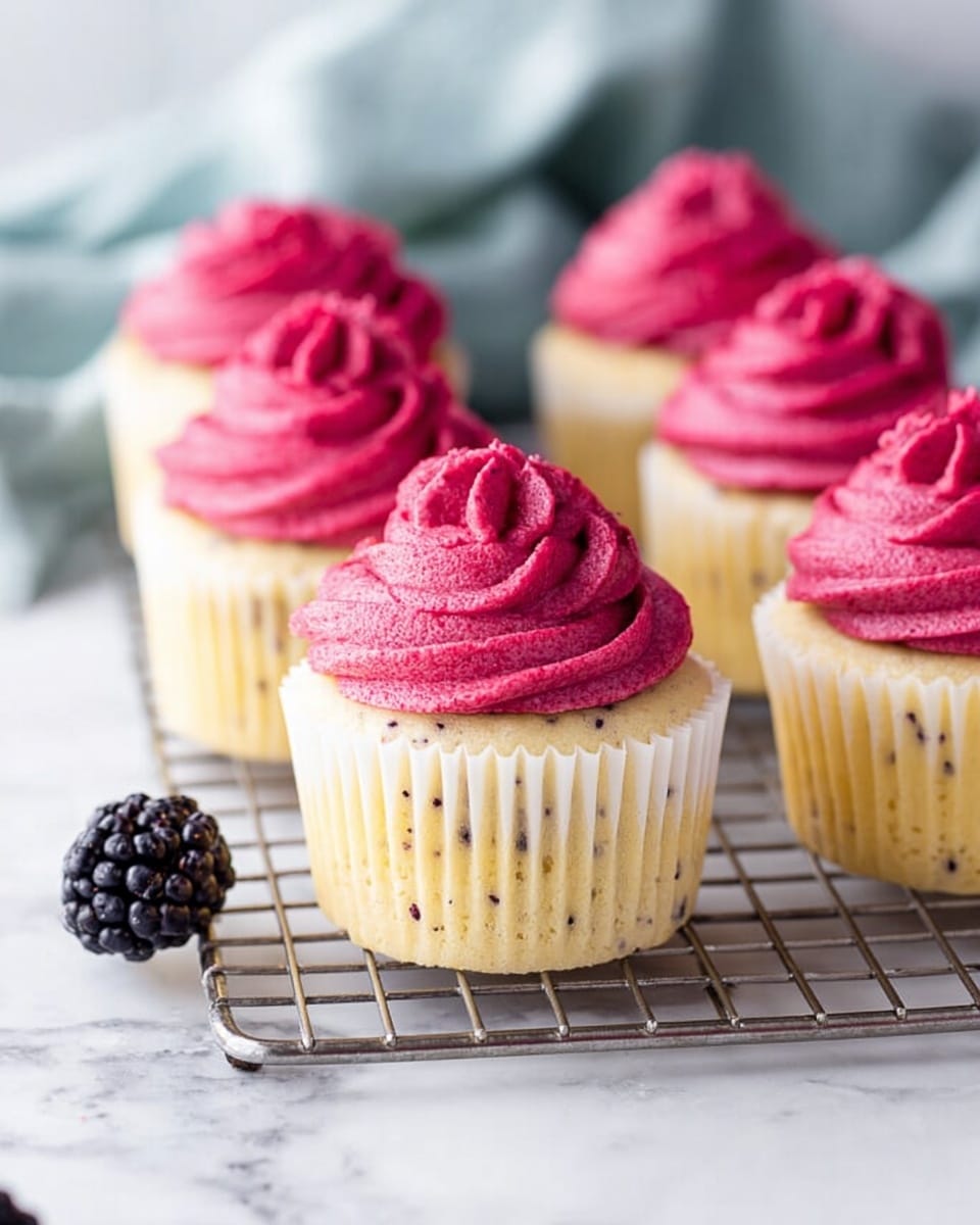 The image shows five cupcakes arranged on a silver cooling rack, placed on a white marbled surface. Each cupcake has two layers: the bottom layer is a light yellow cake with visible small dark specks, wrapped in a white cupcake liner. The top layer is a thick, smooth swirl of bright pink frosting with a slightly shiny texture. A single dark blackberry sits on the white marbled surface near the rack, adding contrast. The background is softly blurred with a hint of light blue cloth. photo taken with an iphone --ar 4:5 --v 7