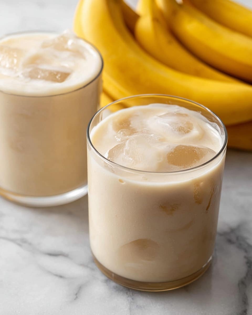 Two clear glasses filled with a creamy, light beige smoothie that has visible ice cubes floating and creating a cool texture. Each glass shows a smooth, slightly frothy top layer with some lighter patches. In the background, there is a bunch of ripe yellow bananas adding a pop of bright color. The glasses are placed on a white marbled surface, enhancing the fresh and clean look of the scene. photo taken with an iphone --ar 4:5 --v 7