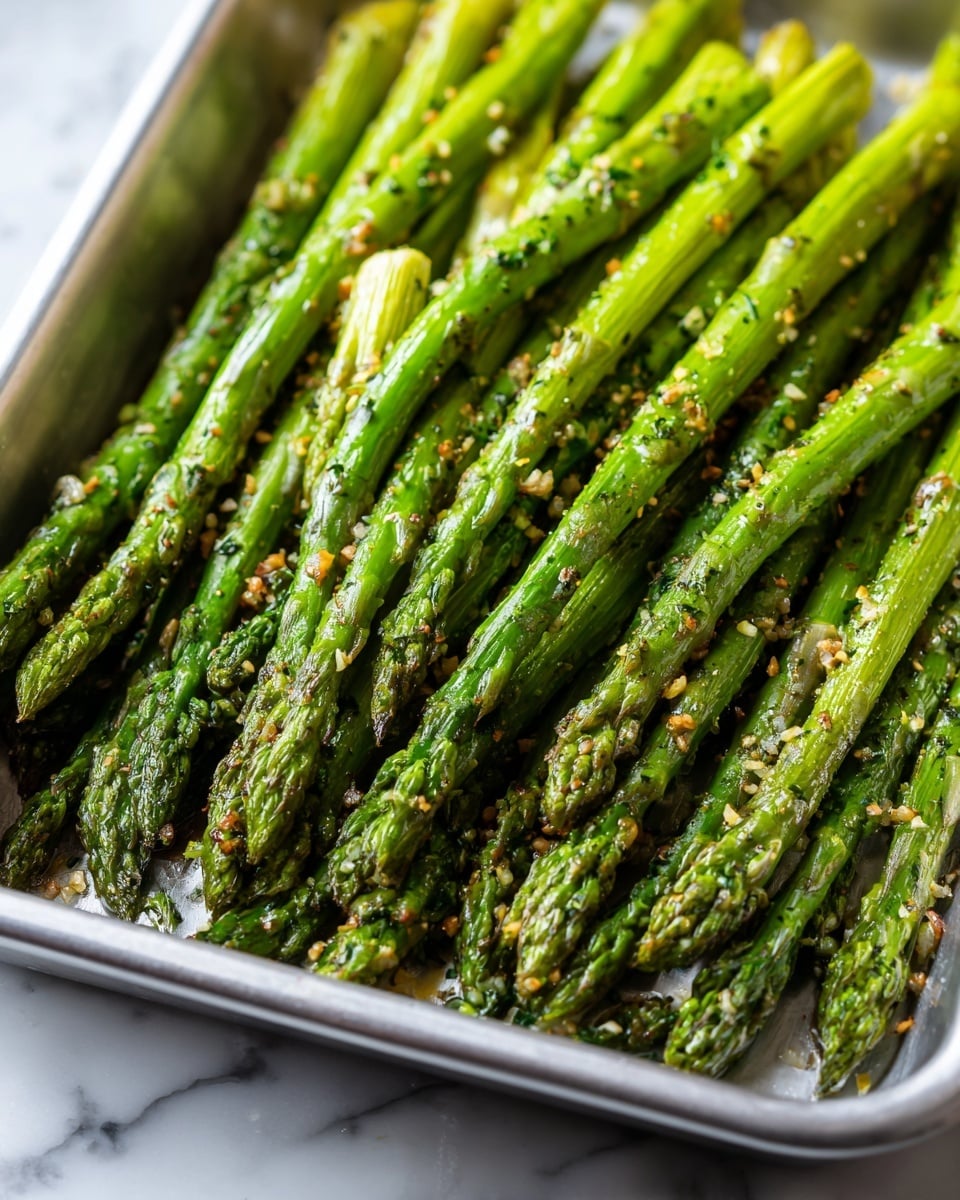 The image shows a metal tray filled with several roasted asparagus spears arranged in parallel rows, each asparagus spear is bright green with slight browning and grill marks, and they are coated in a light layer of oil and herbs with some small bits of chopped green garnish scattered over them; the texture looks tender but still firm. The tray sits on a surface with a white marbled texture. photo taken with an iphone --ar 4:5 --v 7