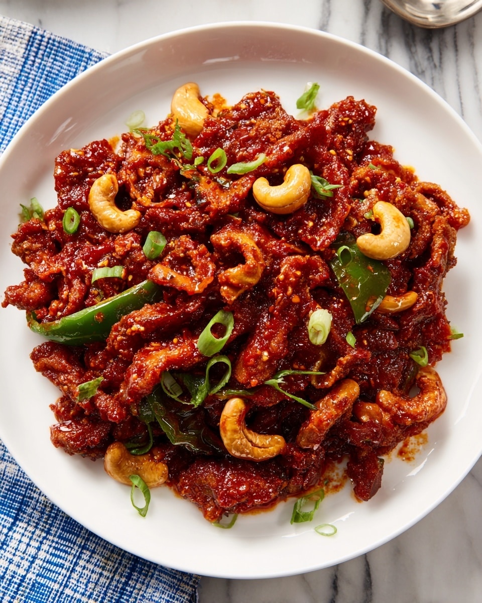 The dish shows a pile of crispy fried strips coated in a thick, glossy red sauce, spread evenly on a white round plate. The strips have a rough, crunchy texture and are mixed with some whole cashews and thin, curved slices of green bell pepper. Small green herb pieces are scattered on top, adding a fresh contrast to the rich red color. The food looks shiny and sticky, with the sauce clinging well to each piece, and the plate sits on a white marbled surface with a folded checkered cloth nearby. photo taken with an iphone --ar 4:5 --v 7