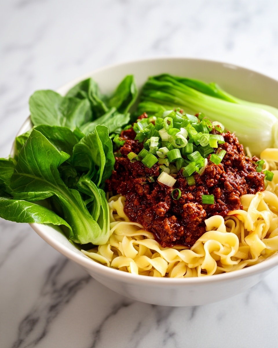 A close-up view of a bowl filled with three main layers: at the bottom are light yellow cooked curly noodles, above them is a thick, dark brown ground meat sauce with a slightly wet texture, and on top is a sprinkle of chopped green onions and crushed peanuts, adding bright green and light brown pops of color. To one side of the bowl, there is a piece of bright green leafy vegetable. The bowl is white, and the background is a white marbled texture. Photo taken with an iphone --ar 4:5 --v 7