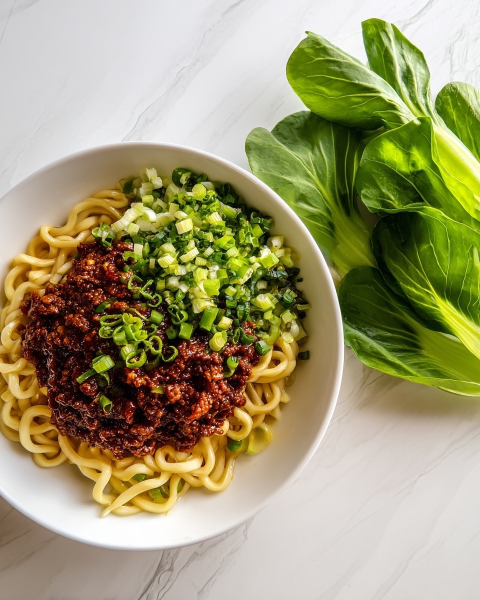 A white bowl on a white marbled surface holds a colorful noodle dish with three main layers. The bottom layer is curly, light yellow noodles, soft in texture. On top of the noodles, there is a thick, dark reddish-brown minced meat sauce with a slightly oily shine. The top layer is chopped green onions, adding a fresh green color and slight crunch. To the side, there is a small bunch of bright green leafy vegetables with smooth leaves. The dish looks fresh and tasty with contrasting colors and textures. Photo taken with an iphone --ar 4:5 --v 7