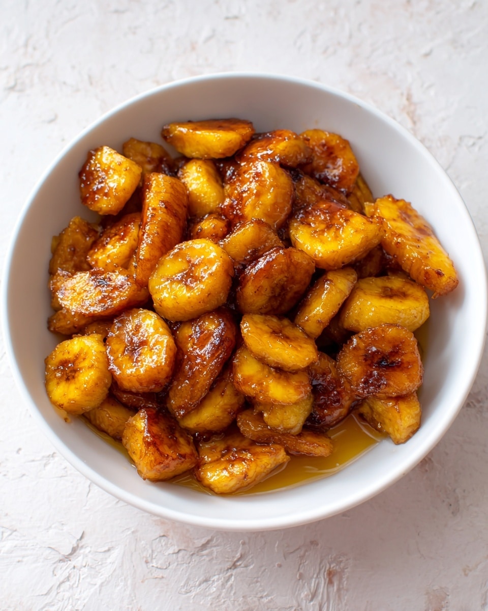 The image shows a white bowl filled with cooked plantain pieces cut into bite-sized chunks. The plantains have a golden brown color with some pieces appearing slightly darker, indicating caramelization. The texture looks soft and slightly sticky, coated with a shiny layer of syrup or sauce that pools gently at the bottom. The background is a white marbled texture. Photo taken with an iphone --ar 4:5 --v 7