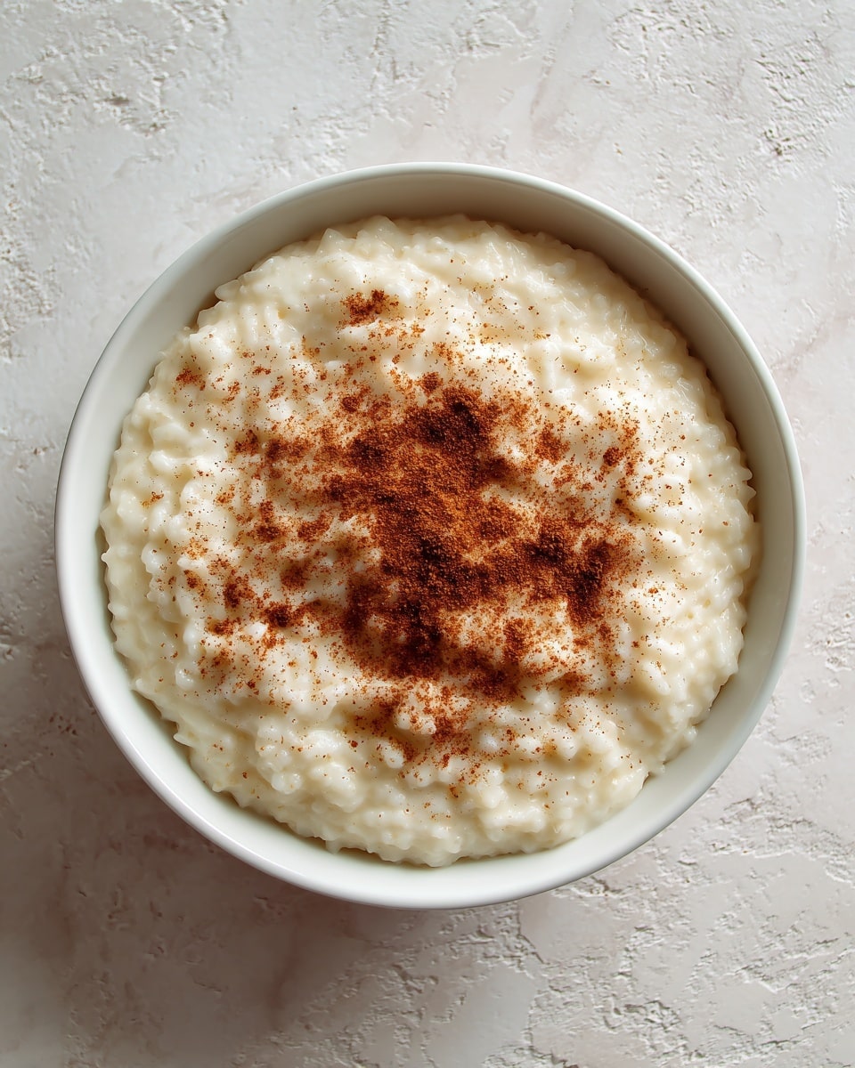 A white bowl filled with creamy rice pudding showing a smooth, slightly lumpy texture on top. The pudding is light cream in color with tiny white rice grains visible, sprinkled with a fine layer of brown cinnamon powder spread over the surface. The bowl sits on a white marbled texture background, and the lighting highlights the soft, moist look of the pudding. photo taken with an iphone --ar 4:5 --v 7