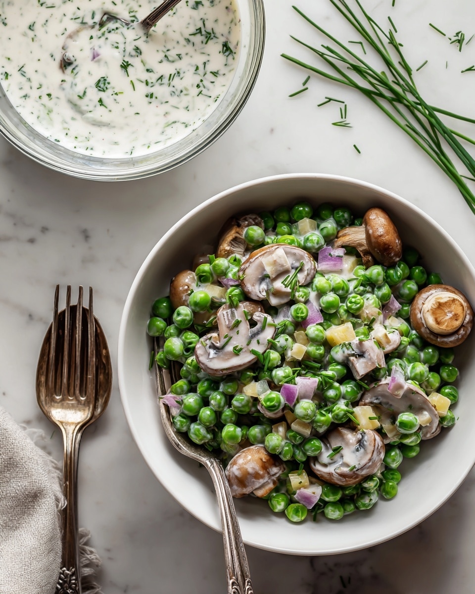 A bowl filled with a creamy salad made of bright green peas covered in a white sauce with small green herb flecks, mixed with sliced brown mushrooms showing their soft texture, and small cubes of yellow cheese along with tiny bits of purple onion scattered throughout. The salad is placed in a white bowl on a white marbled surface, with some fresh chopped green herbs sprinkled nearby. A small glass bowl containing more white sauce with herb bits is visible in the background. photo taken with an iphone --ar 4:5 --v 7