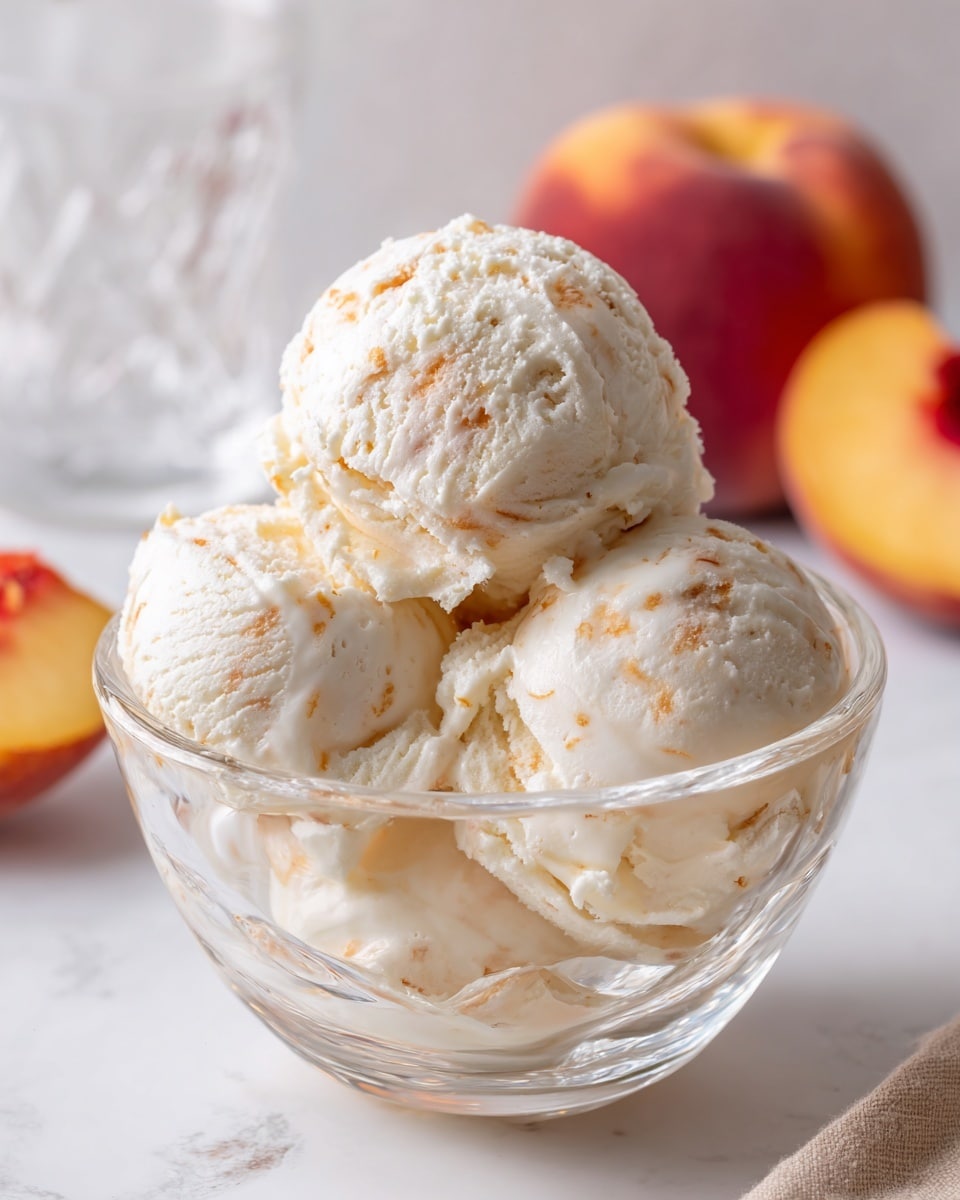 A clear glass dessert bowl filled with three large scoops of creamy peach ice cream, each scoop showing a smooth, slightly textured surface with tiny peach-colored specks spread evenly throughout. The ice cream's pale off-white color is mixed with soft warm peach tones, giving it a fresh and inviting look. The bowl is placed on a white marbled surface, and in the soft-focus background, there are ripe peaches and a glass with a golden drink, adding warm, natural colors to the scene. photo taken with an iphone --ar 4:5 --v 7